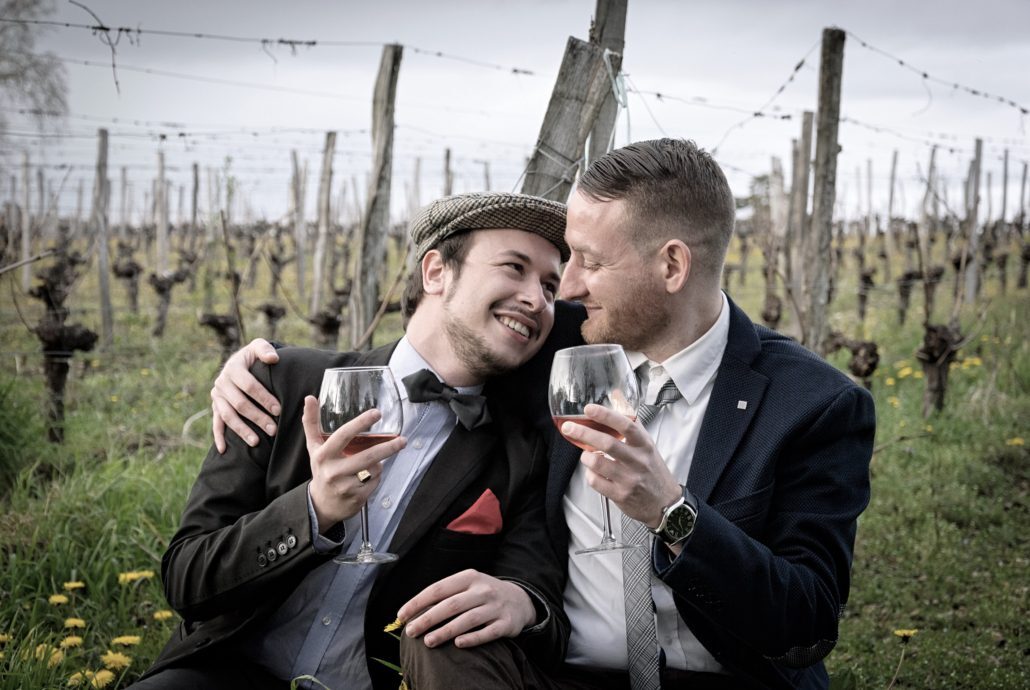 Two men sitting in a vineyard holding wine glassesDescription automatically generated